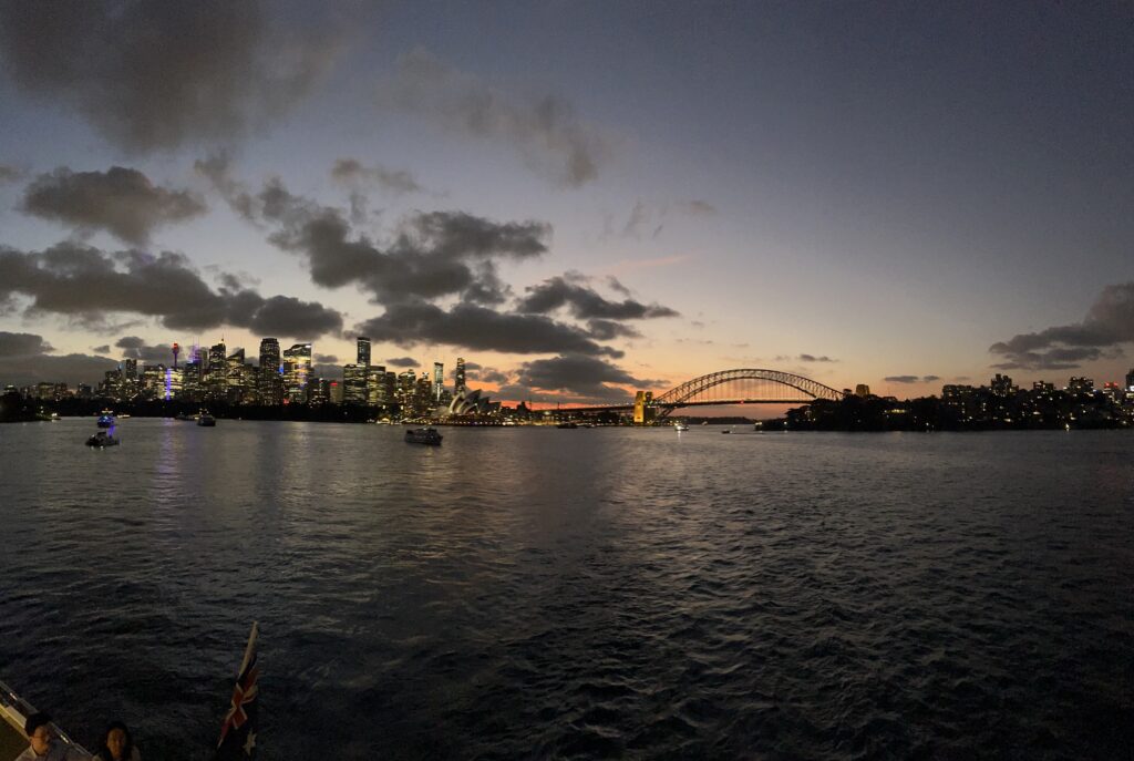 A photo of Sydney Harbour at dusk, from the water look at the sun setting behind the Harbour Bridge and Opera House